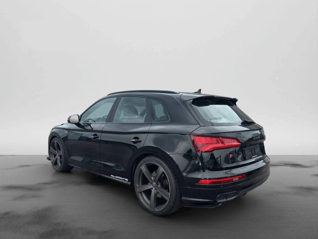 Schwarzer Audi SUV, schräg von hinten links aufgenommen. Das Fahrzeug steht auf nassem Asphalt vor neutralem, grauem Hintergrund und wirkt sportlich mit roten Rückleuchten und grossen Felgen.
